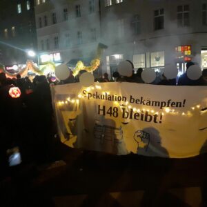 Bewohner:innen der H48 halten ein mit einer Lichterkette dekoriertes Transparent mit der Aufschrift "Spekulation bekämpfen! H48 bleibt", im Hintergrund sieht man weitere Demonstrierende und den Kiezdrachen